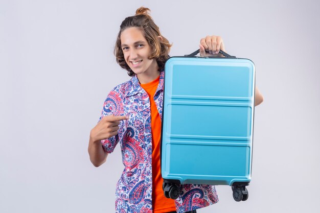 Uomo giovane viaggiatore con la valigia blu guardando fiducioso sorridente con la faccia felice pronta a viaggiare in piedi su sfondo bianco