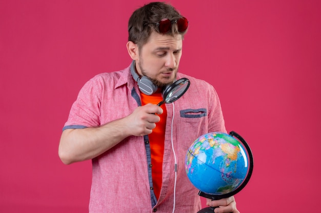 Uomo giovane viaggiatore con le cuffie guardando il globo attraverso la lente di ingrandimento in piedi su sfondo rosa