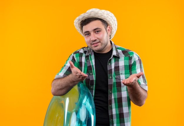 Uomo giovane viaggiatore in camicia a quadri e cappello estivo che tiene anello gonfiabile con le braccia alzate come fare domanda in piedi sopra la parete arancione