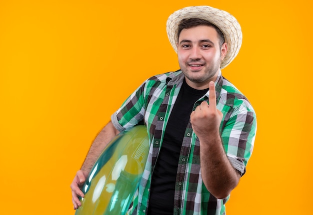 Uomo giovane viaggiatore in camicia a quadri e cappello estivo che tiene anello gonfiabile sorridente che mostra il dito indice in piedi sopra la parete arancione