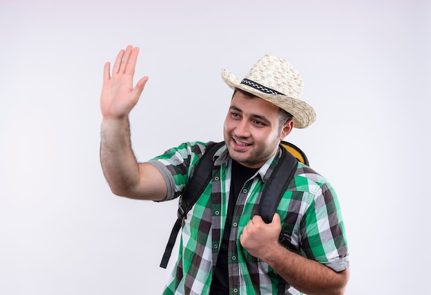 Uomo giovane viaggiatore in camicia a quadri e cappello estivo con zaino che sorride allegramente agitando con la mano in piedi sopra il muro bianco