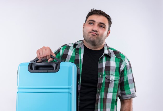 Uomo giovane viaggiatore in camicia controllata che tiene la valigia che osserva da parte con l'espressione triste sul viso in piedi sopra il muro bianco