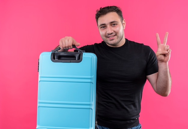 Uomo giovane viaggiatore in maglietta nera che tiene la valigia felice e positiva con il sorriso sul viso che mostra il segno di vittoria in piedi sopra il muro rosa