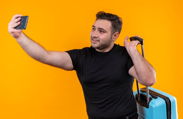 Uomo giovane viaggiatore in maglietta nera che tiene la valigia prendendo selfie, utilizzando il suo smartphone sorridendo alla telecamera in piedi sopra la parete arancione
