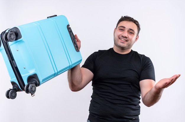 Uomo giovane viaggiatore in maglietta nera che tiene la valigia sorridente allegramente diffondendo con il braccio di lato in piedi sopra il muro bianco