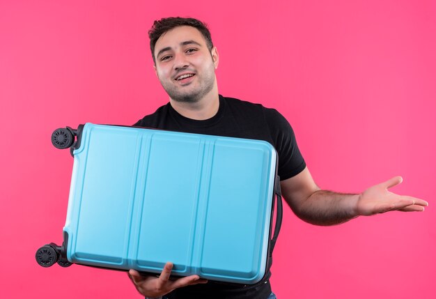 Uomo giovane viaggiatore in maglietta nera che tiene la valigia sorridente amichevole in piedi sopra il muro rosa