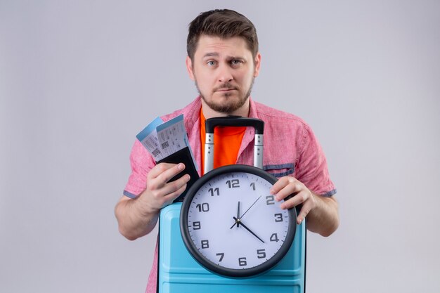 Uomo giovane viaggiatore in possesso di biglietti aerei e valigia con orologio che guarda l'obbiettivo con seria espressione fiduciosa sul viso in piedi su sfondo bianco isolato