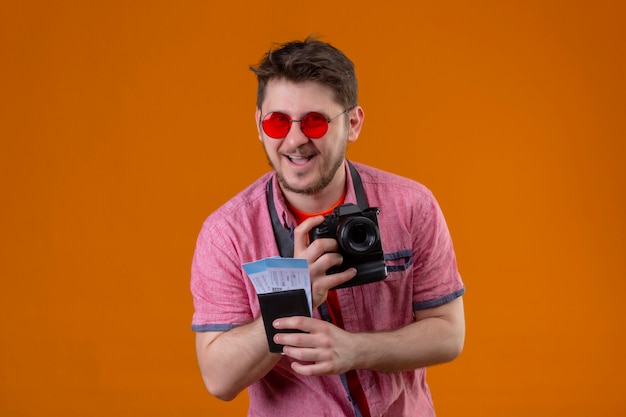 Uomo giovane viaggiatore indossando occhiali da sole con fotocamera in possesso di biglietti aerei che guarda l'obbiettivo felice ed è uscito sorridendo allegramente in piedi su sfondo arancione