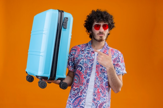 Uomo giovane viaggiatore riccio bello confuso che indossa occhiali da sole e che tiene la valigia indicandolo sullo spazio arancione isolato con lo spazio della copia