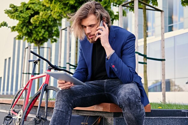 Uomo hipster con lunghi capelli biondi parla tramite smartphone e tiene tablet PC con bicicletta a velocità singola sullo sfondo.