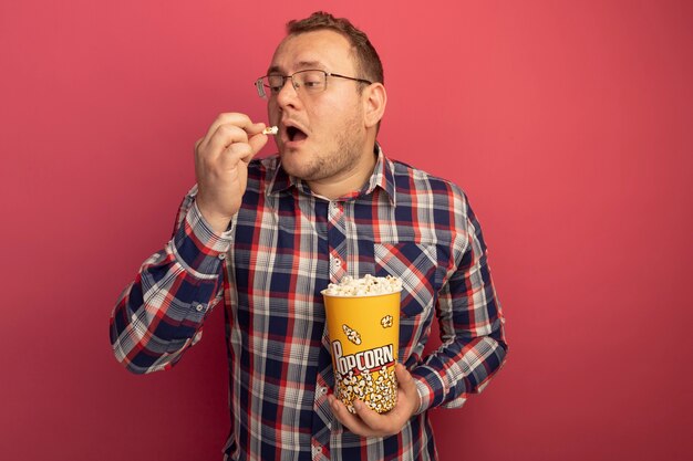 Uomo in bicchieri e camicia a quadri che tiene secchio con popcorn mangiare con piacere in piedi sopra il muro rosa