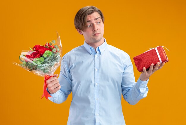 uomo in blueshirt holding bouquet di rose rosse e presente cercando confuso avendo dubbi il concetto di San Valentino in piedi sopra la parete arancione