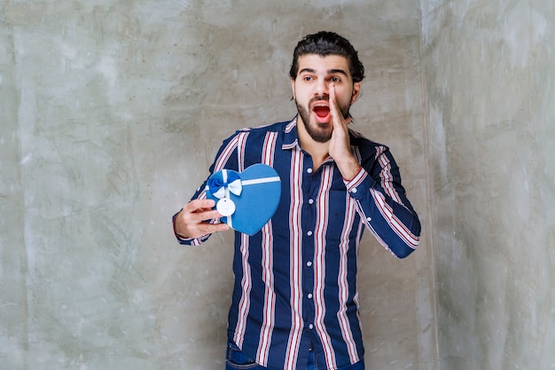 Uomo in camicia a righe che tiene una scatola regalo blu a forma di cuore e chiama qualcuno a venire nelle vicinanze