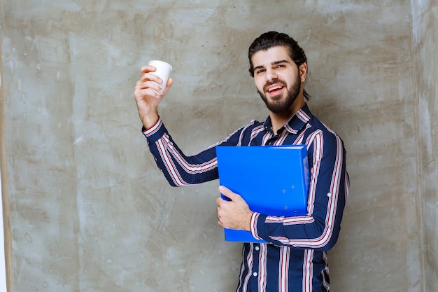 Uomo in camicia a righe con in mano una cartellina blu e una tazza bianca usa e getta