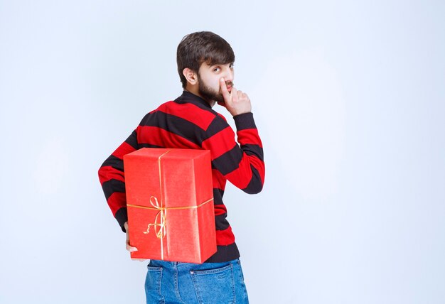 Uomo in camicia a righe rossa che nasconde una confezione regalo rossa dietro di sé.