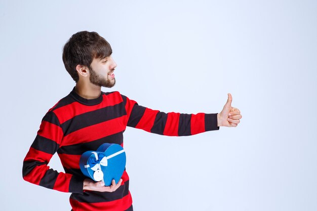 Uomo in camicia a righe rossa che tiene una scatola regalo blu a forma di cuore e mostra il pollice in alto segno.