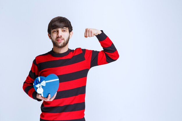 Uomo in camicia a righe rosse che tiene una scatola regalo blu a forma di cuore e mostra il pugno.