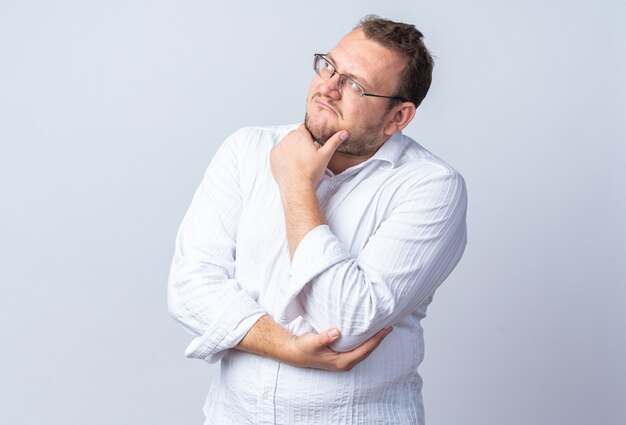 Uomo in camicia bianca con gli occhiali che guarda da parte con la mano sul mento pensando in piedi sul muro bianco