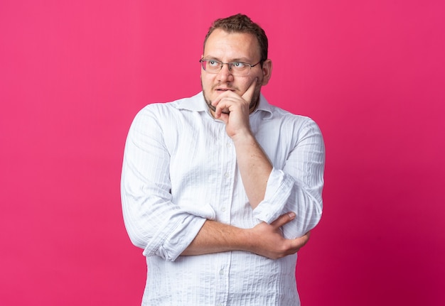 Uomo in camicia bianca con gli occhiali che guarda da parte con un'espressione pensierosa sul viso pensando in piedi sul muro rosa