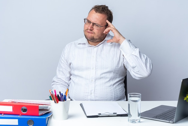 Uomo in camicia bianca con gli occhiali che guarda da parte confuso e molto ansioso seduto al tavolo con cartelle per ufficio portatile e appunti sul muro bianco che lavora in ufficio
