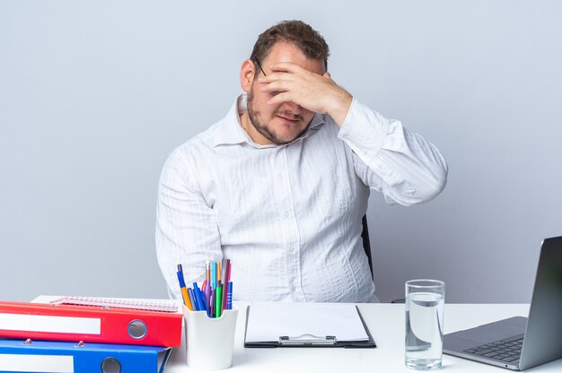 Uomo in camicia bianca con gli occhiali che sembra infastidito e stanco cono gli occhi con la mano seduto al tavolo con le cartelle dell'ufficio del computer portatile e appunti su bianco