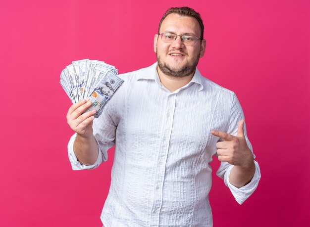 Uomo in camicia bianca con gli occhiali in possesso di contanti puntando con il dito indice ai soldi sorridendo allegramente in piedi sul muro rosa