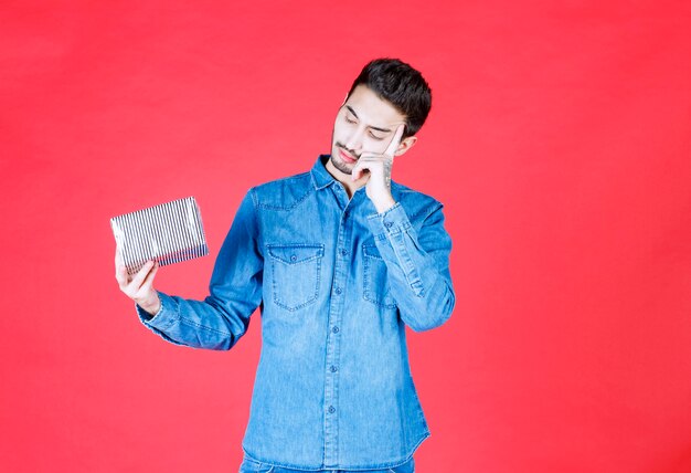 Uomo in camicia di jeans che tiene in mano una confezione regalo d'argento e sembra confuso e inaspettato.