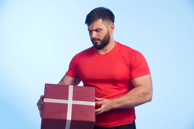 Uomo in camicia rossa che tiene una grande confezione regalo e sembra serio.