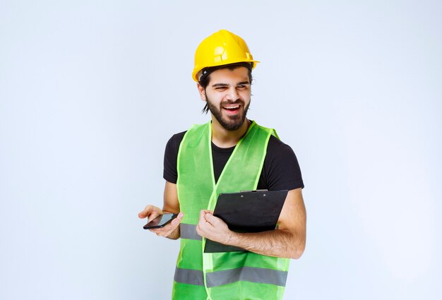 Uomo in casco giallo che tiene una cartella di progetto e uno smartphone.