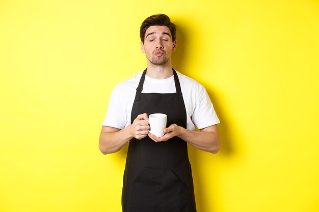 Uomo in grembiule nero che porta una tazza di caffè e aspetta un bacio, in piedi su sfondo giallo.