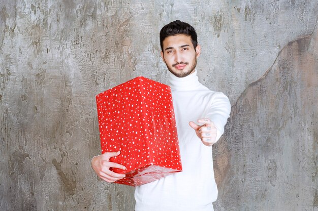 Uomo in maglione bianco che tiene una confezione regalo rossa con puntini bianchi su di esso e che mostra un segno positivo della mano.