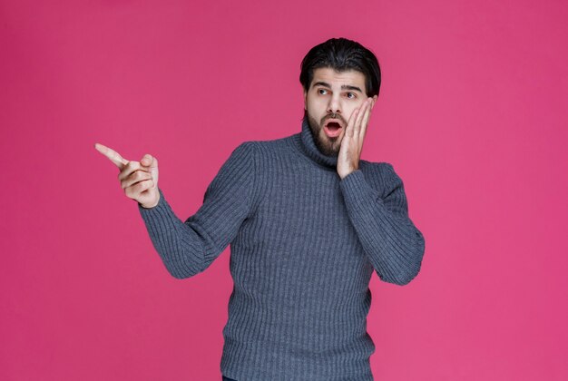 Uomo in maglione grigio che punta a qualcosa con sorpresa e si tiene la testa.