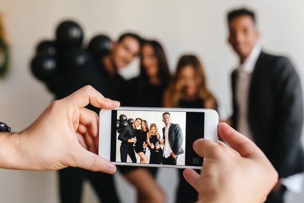 Uomo in orologio da polso che tiene smartphone bianco e sta andando a fare foto di amici che si divertono alla festa