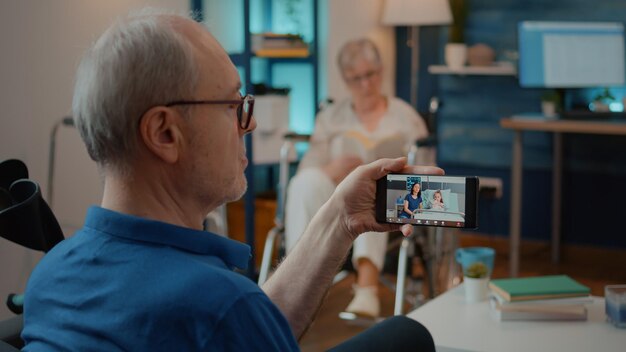 Uomo in pensione che parla con la figlia e la nipote in ospedale in videochiamata, seduto accanto alle stampelle. Persona che utilizza lo smartphone per chattare in videoconferenza online con i parenti in clinica.