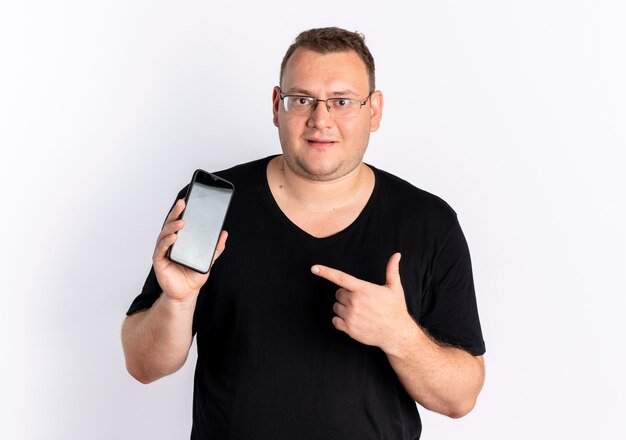 Uomo in sovrappeso che indossa la maglietta nera con gli occhiali che mostra lo smartphone che punta con il dito guardando fiducioso in piedi sopra il muro bianco