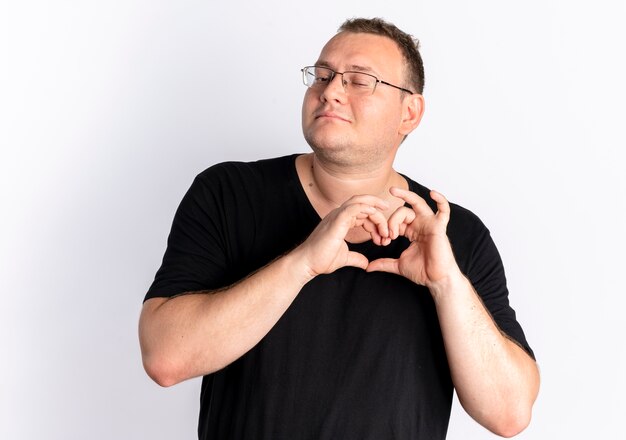Uomo in sovrappeso con gli occhiali che indossa la maglietta nera che fa il gesto del cuore con le dita che provano emozioni positive in piedi sopra il muro bianco
