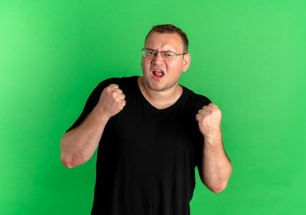 Uomo in sovrappeso con gli occhiali che indossa la maglietta nera che stringe i pugni felici ed eccitati in piedi sopra il muro verde