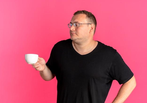 Uomo in sovrappeso con gli occhiali che indossa la maglietta nera che tiene la tazza di caffè che osserva da parte con la faccia seria sopra il rosa