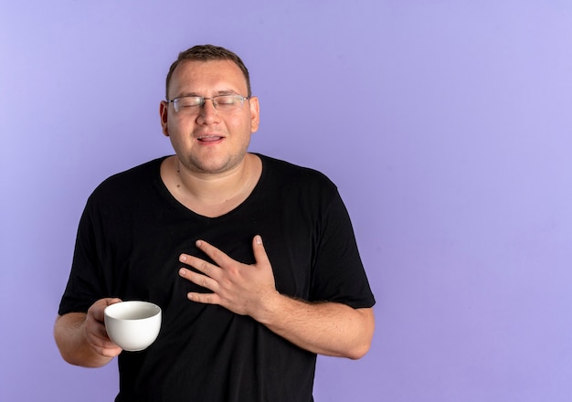Uomo in sovrappeso con gli occhiali che indossa la maglietta nera che tiene la tazza di caffè che tiene la mano sul petto sentendosi grato in piedi sopra il muro blu