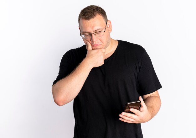 Uomo in sovrappeso con gli occhiali che indossa la maglietta nera guardando lo schermo del suo smartphone perplesso in piedi sopra il muro bianco