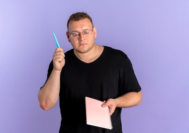 Uomo in sovrappeso con gli occhiali che indossa t-shirt nera in possesso di taccuino e penna che sembra perplesso in piedi sopra la parete blu