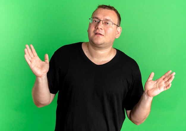 Uomo in sovrappeso con gli occhiali che indossa una maglietta nera che sembra confuso e incerto allargando le braccia ai lati senza risposta sul verde