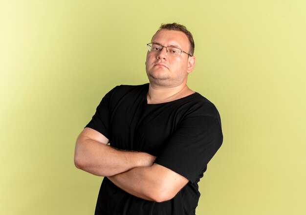 Uomo in sovrappeso con gli occhiali che indossa una maglietta nera con seriosu espressione sicura con le braccia incrociate sul petto in piedi sopra la parete chiara