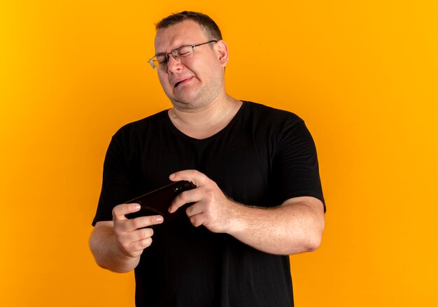 Uomo in sovrappeso con gli occhiali che indossa una maglietta nera guardando lo schermo dello smartphone che sembra scontento in piedi sopra il muro arancione