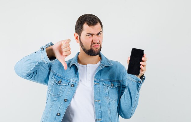 Uomo in stile retrò che mostra il pollice verso il basso mentre mostra il telefono in giacca, t-shirt e sembra insoddisfatto. vista frontale.