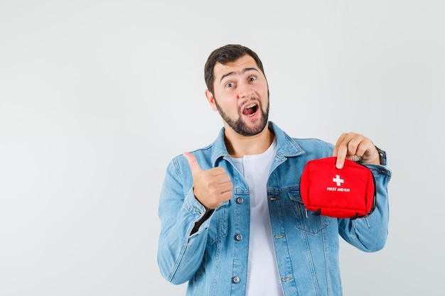 Uomo in stile retrò che tiene il kit di pronto soccorso mentre mostra il pollice in giacca, maglietta e sembra ottimista. vista frontale.