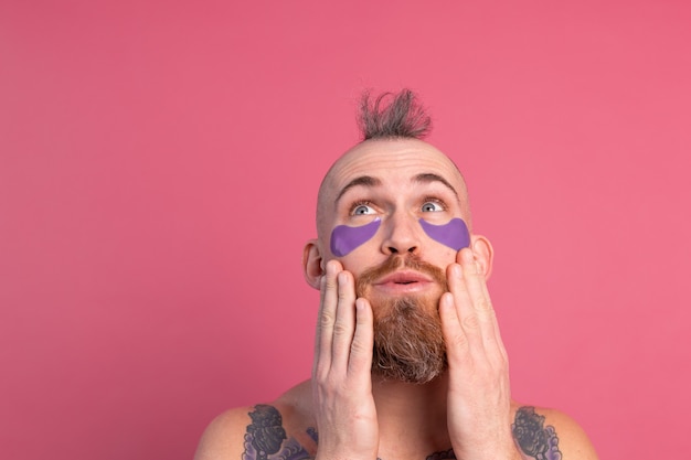 Uomo in topless tatuato bello barbuto europeo con maschera di bende sull'occhio viola in posa per la fotocamera sul rosa