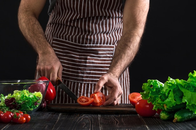 Uomo in un grembiule a righe che prepara insalata su un tavolo di legno. Le mani degli uomini tagliano il pomodoro per fare un'insalata su sfondo nero. Concetto di cibo sano