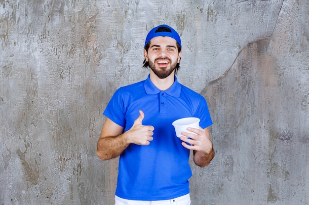 Uomo in uniforme blu che tiene in mano un bicchiere di plastica da asporto e mostra un segno positivo con la mano.