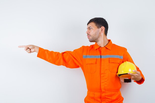 Uomo industriale in uniforme che tiene il casco, indicando il lato e guardando concentrato, vista frontale.